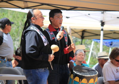 Two men perform with a drum under tents at an outdoor festival.