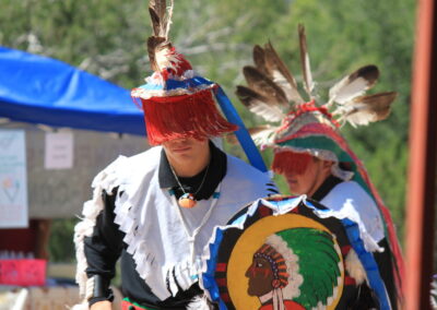 Two people in Native American regalia with feathered headdresses and a decorated shield.