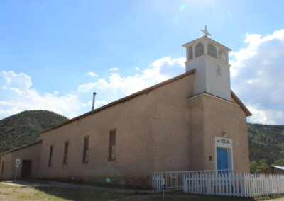 Small beige church with a blue door and white fence, hills in the background.