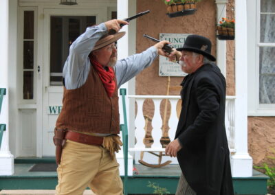 Two men in Western attire on a porch, one pointing a pistol at the other.