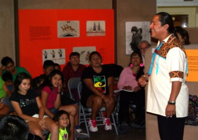 Speaker in traditional attire addresses a seated audience of children and adults.