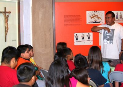 Man leads a discussion with seated children beside an orange exhibit wall.