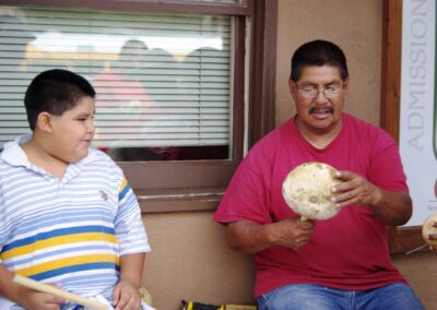 Man in a red shirt shows a shell to a striped-shirt boy outdoors.