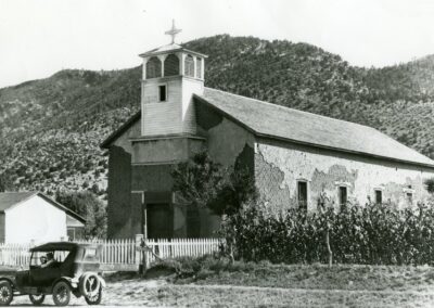 Old church with a small bell tower and a vintage car in front of hills.