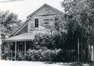 Old wooden house with peeling siding, partially obscured by overgrown bushes.