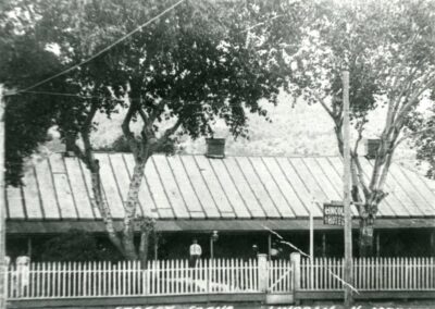 Old B&W photo of a house with a metal roof, white picket fence, and trees.
