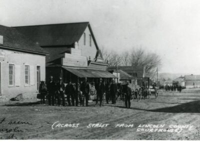 Historic street scene with storefronts and a crowd gathered outside a large building.