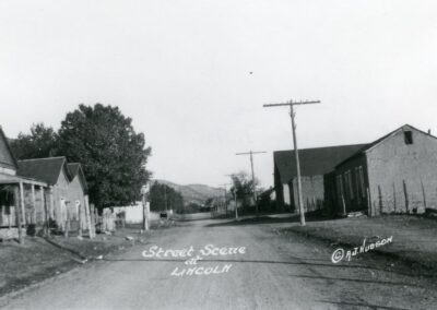 Black-and-white street scene in a small town with houses and utility poles along a dirt road.