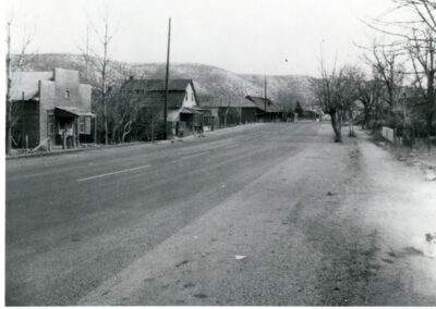 Black-and-white street scene in a small town with houses, trees, and distant hills.
