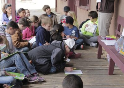Children sit on a porch writing while a teacher stands nearby.