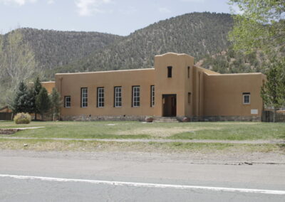 Tan adobe-style single-story building beside a road with mountains in the background.