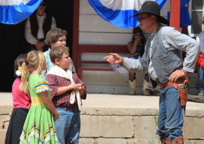 Cowboy in hat and chaps speaks to children on a stage with blue banners.
