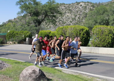 Group of runners jogging on a sunny road beside green bushes and hills.