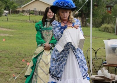 Two women in Victorian-era dresses outdoors, one in blue and white with a blue hat.