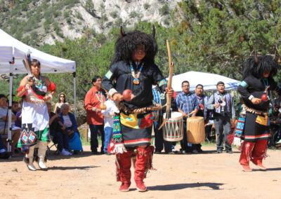 Dancers in black regalia with red pants perform outdoors as drummers and spectators watch.