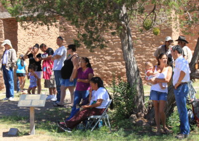 Group of people gathered outdoors near a brick wall and a tree, some standing, some seated.